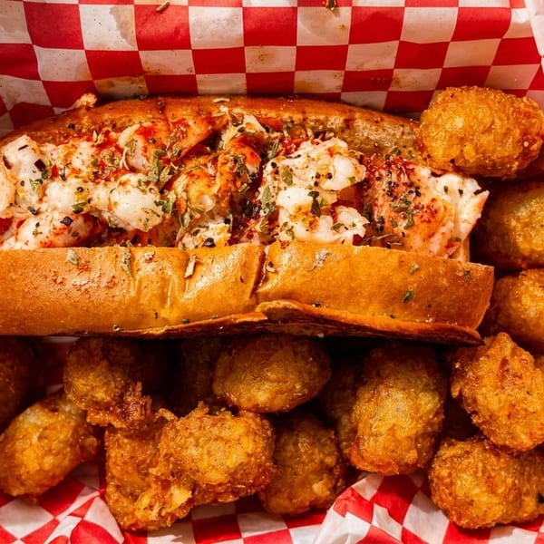 Lobster roll with tater tots in a red and white checkered paper-lined basket.