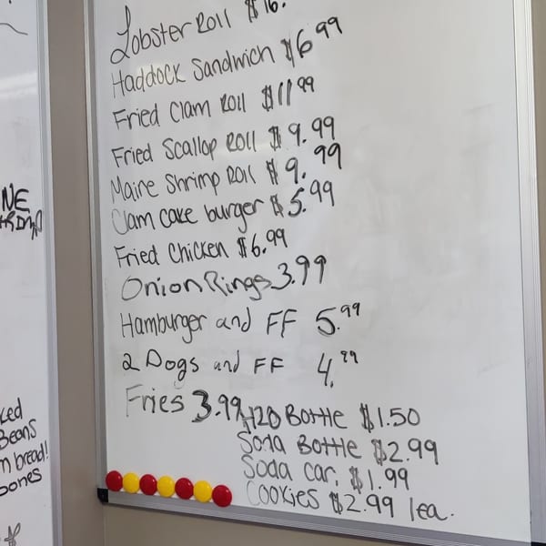 A handwritten menu on a whiteboard lists sandwiches, sides, and drinks with prices.