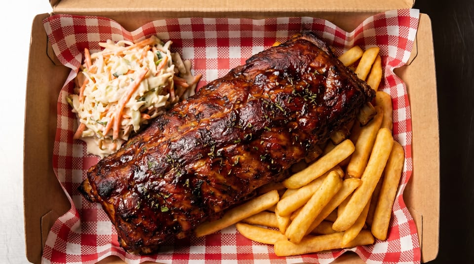 BBQ ribs with coleslaw and french fries in a takeout box with red and white checkered paper.